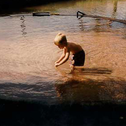A shirtless blonde male child, dressed in dark shorts plays in a freshwater pond at Moab Springs Ranch.