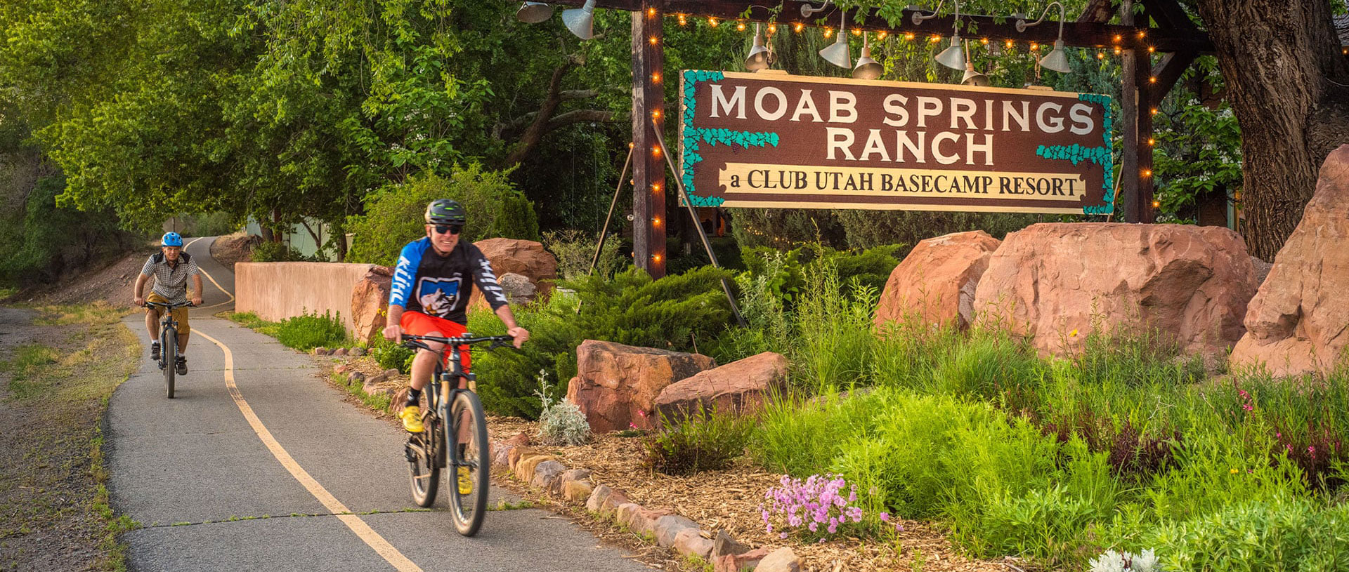 Two male bicyclists ride past canopies of green leaves from tall cottonwood trees, multi colored flora, large sandstone boulders and the rectangular Moab Springs Ranch sign.
