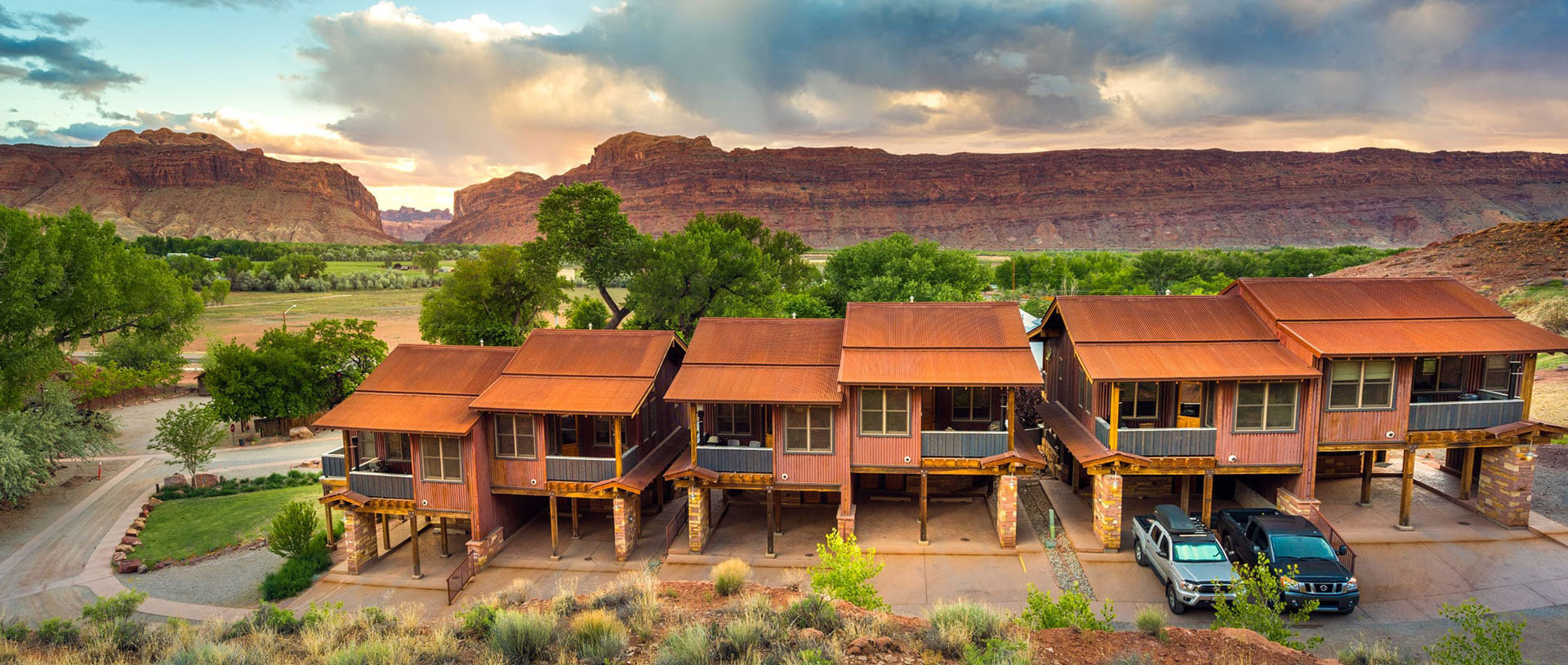 Six brown bungalow units at Moab Springs Ranch stand in a row side by side against a backdrop of lush green cottonwood trees and copper brown ragged cliffs.