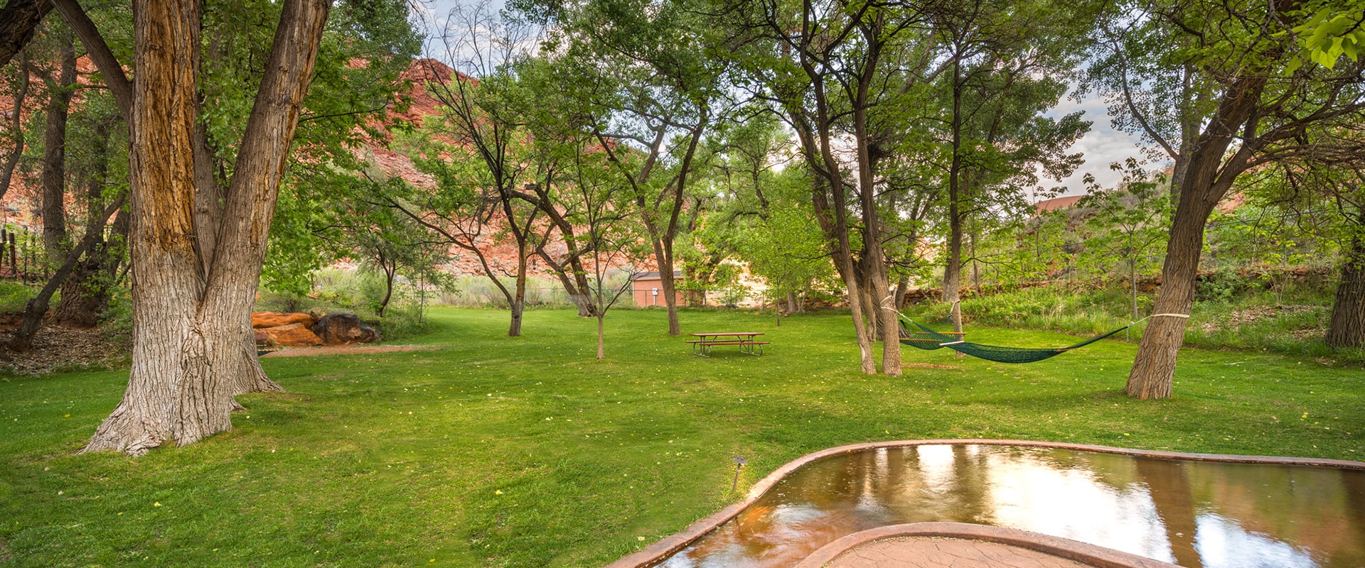 A man-made stream runs across the parkspace of Moab Springs Ranch with towering green trees, a hammock and a picnic table in the background.