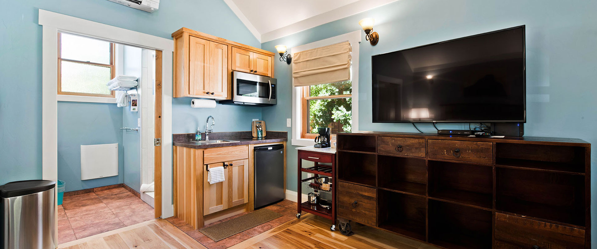The sitting area of Unit #18 at Moab Springs Ranch is furnished with two grey fabric covered armchairs, a floor lamp, two red leather ottomans, and a wall mounted flatscreen TV.