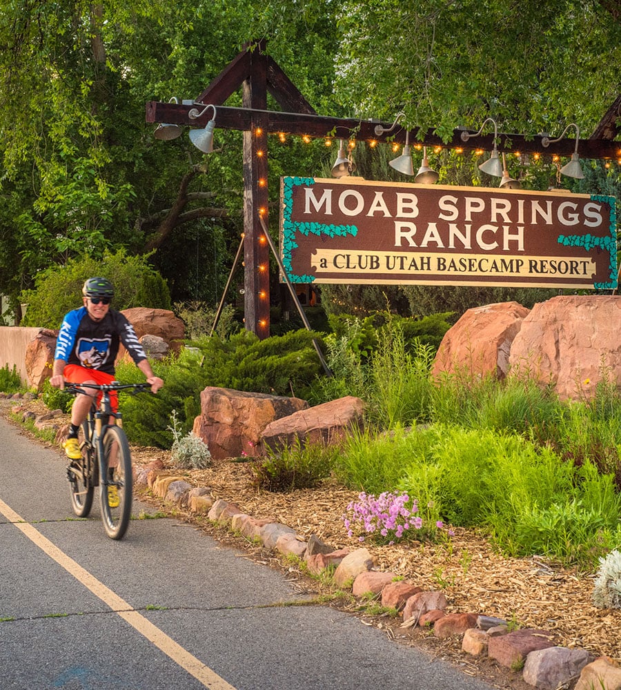 A smiling male bicyclist, wearing a helmet and dark sunglasses, rides along the bike path passing the Moab Springs Ranch sign, large boulders, cobble rocks and green flora and fauna.
