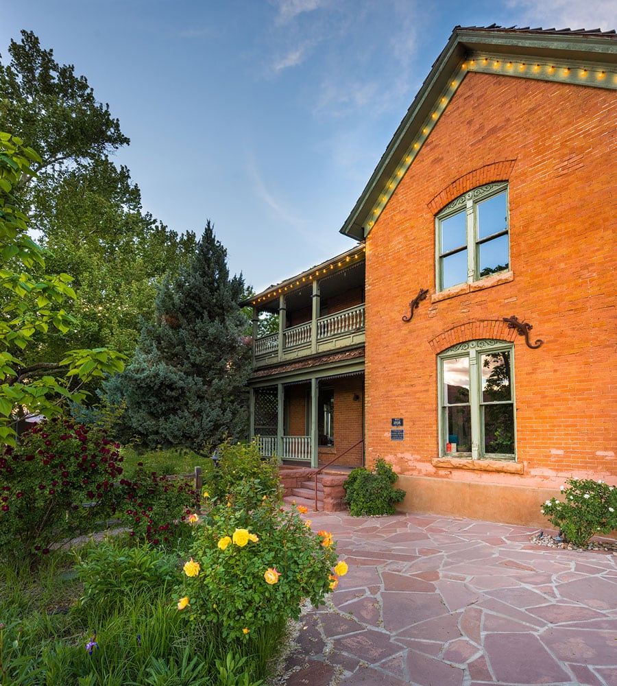 The Main House at Moab Springs Ranch is a two story brown brick structure that sits amongst lush tall trees with multi-colored vegetation at its' doorstep.