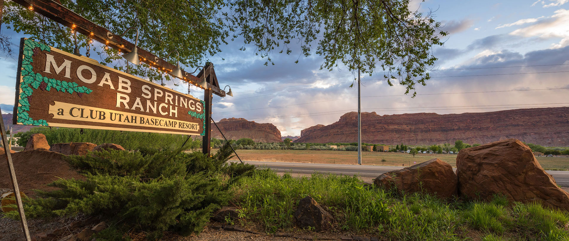 Roadside sign of Moab Springs Ranch in white letter typeset against a brown wood backdrop and green leaf motifs of the corporate logo.